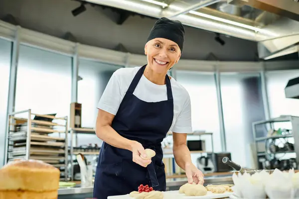 Chef maduro atraente no toque azul sorrindo para a câmera enquanto trabalhava com massa, confeitaria — Fotografia de Stock