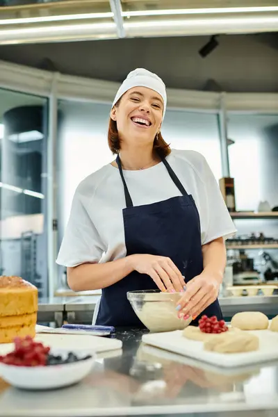 Joyeuse jeune femme attrayante en toque et tablier de travail avec de la pâte et souriant à la caméra — Photo de stock