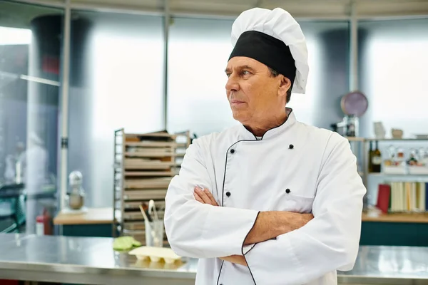 Guapo cocinero jefe maduro en sombrero blanco posando con los brazos cruzados en el pecho y mirando hacia otro lado - foto de stock