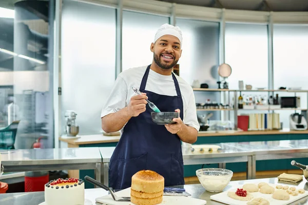 Atractivo cocinero afroamericano alegre en delantal usando cepillo de silicona y sonriendo a la cámara - foto de stock