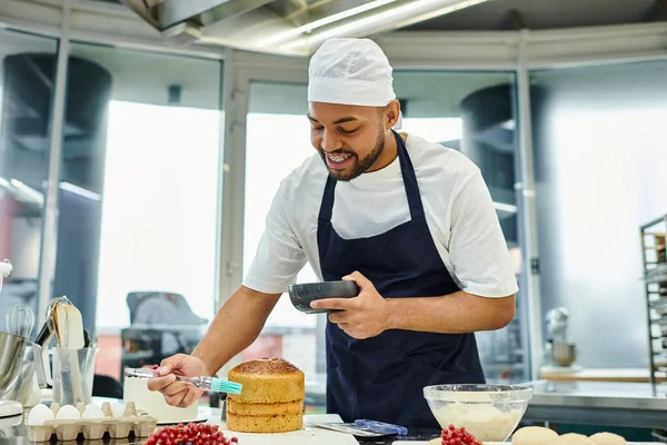 Jolly handsome african american chef in blue toque and apron using silicone brush on delicious cake — Stock Photo