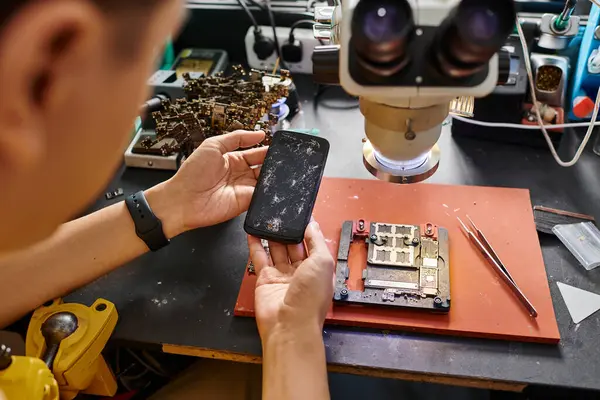 Vista ritagliata di tecnico esperto che tiene il telefono cellulare con schermo rotto vicino alle attrezzature di riparazione — Foto stock