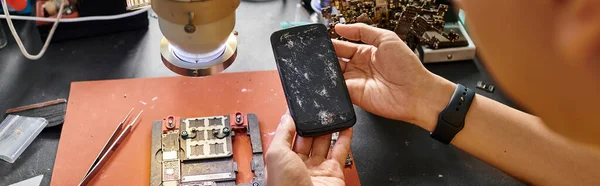 Vista ritagliata di riparatore esperto che tiene il telefono cellulare con schermo rotto vicino ai dispositivi, banner — Foto stock