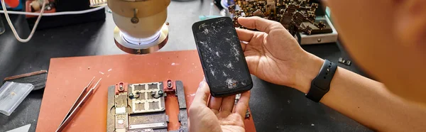 Cropped view of skilled repairman holding mobile phone with broken screen near devices, banner — Stock Photo