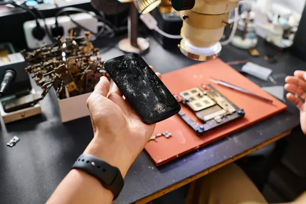 Ausgeschnittene Ansicht eines Werkstattmitarbeiters, der ein Smartphone mit kaputtem Bildschirm in der Nähe elektronischer Werkzeuge in der Werkstatt hält — Stockfoto
