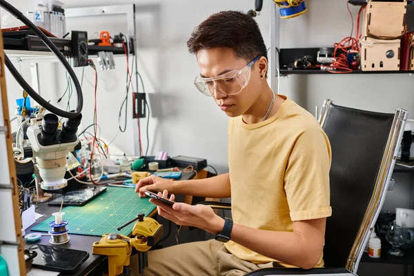 Technicien asiatique en lunettes regardant le téléphone mobile pendant les diagnostics sur le lieu de travail dans l'atelier — Photo de stock