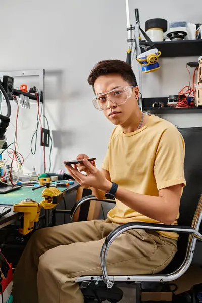 Professional asian repairman in goggles holding smartphone and looking at camera in repair workshop — Stock Photo
