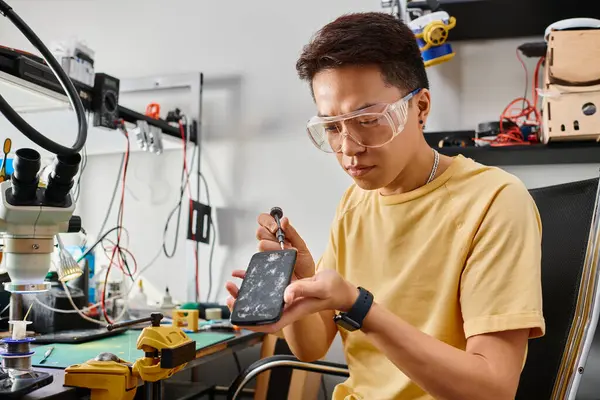 Asian technician in goggles opening smartphone with broken screen with screwdriver in repair shop — Stock Photo