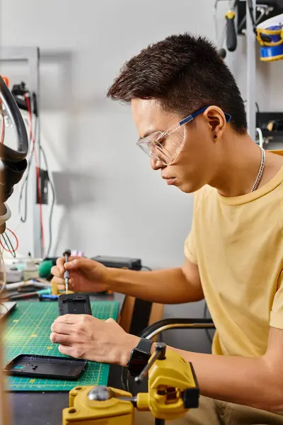 Vue latérale de l'asiatique de réparateur dans des lunettes avec tournevis montage téléphone mobile dans son propre atelier — Photo de stock