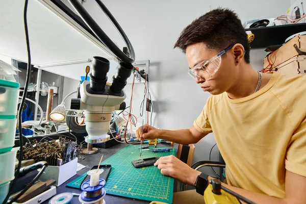 Konzentrierter asiatischer Reparateur mit Brille, der in der Werkstatt technisches Know-how aus Mobiltelefonen herstellt — Stockfoto
