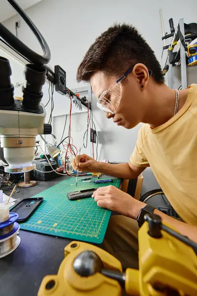 Vue latérale du technicien asiatique dans les lunettes de faire des diagnostics techniques de smartphone dans l'atelier de réparation — Photo de stock