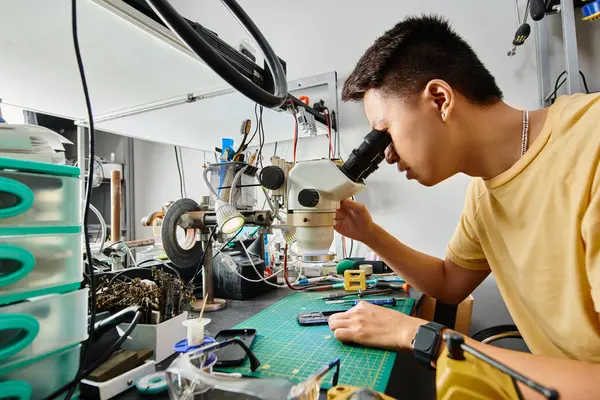 Side view of skilled asian technician looking in microscope while working with electronic equipment — Stock Photo