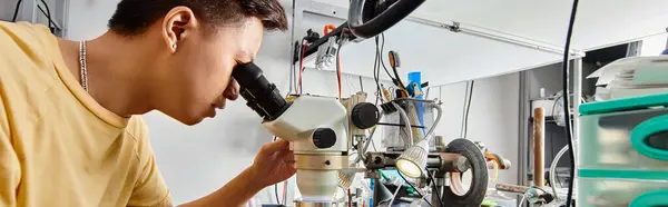 Réparateur asiatique expérimenté regardant au microscope et testant des appareils électroniques, vue latérale, bannière — Photo de stock