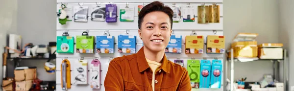 Улыбающийся азиатский продавец, смотрящий на камеру возле прилавка частного магазина электроники, баннер — стоковое фото