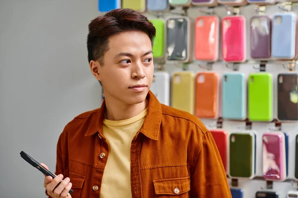 Hombre asiático serio sosteniendo el teléfono móvil y mirando hacia otro lado en la tienda de electrónica privada, pequeña empresa - foto de stock