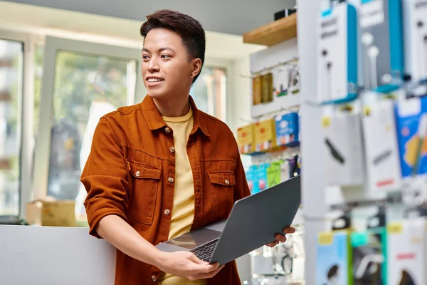 Усміхнений азіатський продавець з сучасним ноутбуком, який дивиться далеко в приватному магазині електроніки, малий бізнес — стокове фото