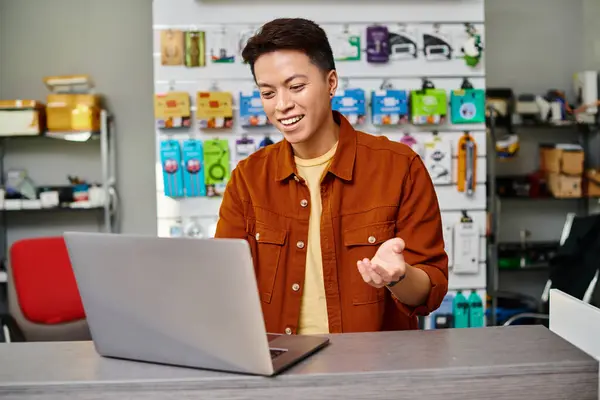 Positive asian salesman gesturing during online sale on laptop on counter in own electronics store — Stock Photo