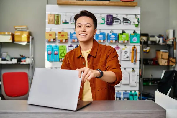 Alegre asiático electrónica tienda propietario mirando cámara cerca de portátil en contador en privado tienda - foto de stock