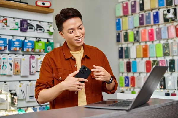 Souriant asiatique entrepreneur montrant smartphone lors d'un appel vidéo sur ordinateur portable en magasin privé — Photo de stock