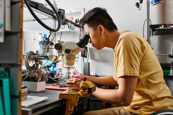 Vista lateral do profissional asiático especialista com microscópio examinando dispositivo eletrônico na oficina — Fotografia de Stock