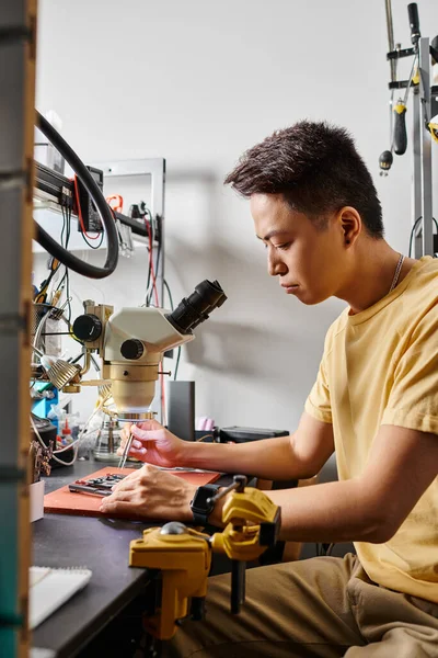 Опытный азиатский ремонтник, работающий с микроскопом и гаджетами в мастерской, малый бизнес — стоковое фото