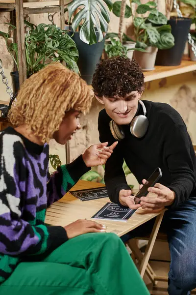 Heureux couple interracial avec cheveux bouclés choisir menu sur smartphone dans un café végétalien rempli de plantes — Photo de stock