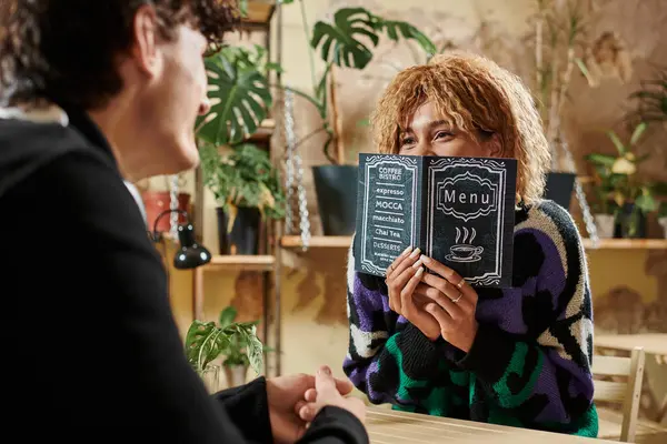 Happy african american woman covering face while holding menu and looking at boyfriend in vegan cafe — Stock Photo