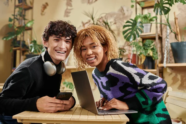 Pareja diversa positiva utilizando dispositivos mientras están sentados juntos en la mesa en la cafetería vegana llena de plantas - foto de stock