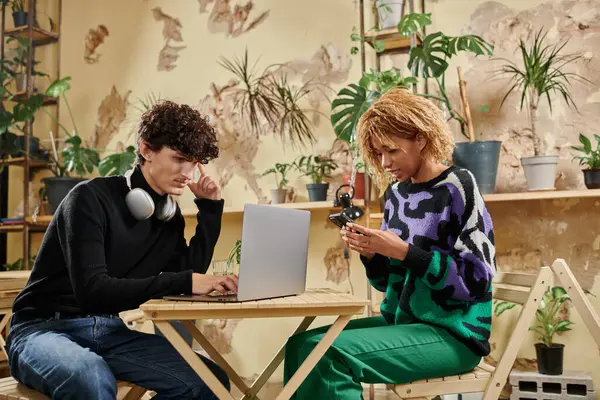 Jovem e diverso casal usando dispositivos enquanto sentado toegther à mesa no café vegan repleto de plantas — Fotografia de Stock