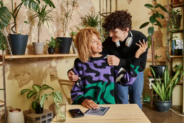 Curly young man hugging his african american girlfriend sitting near menu on table of vegan cafe — Stock Photo