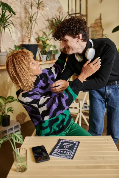 Joven rizado abrazando a su novia afroamericana sentada cerca del menú en la mesa de café vegano - foto de stock