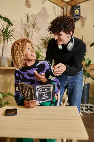 Positivo y joven hombre y mujer en el menú de lectura de ropa casual en la cafetería vegana, pareja interracial - foto de stock