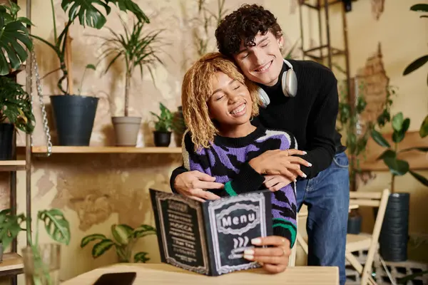 Affectionate and curly young man embracing african american girlfriend near menu in vegan cafe — Stock Photo