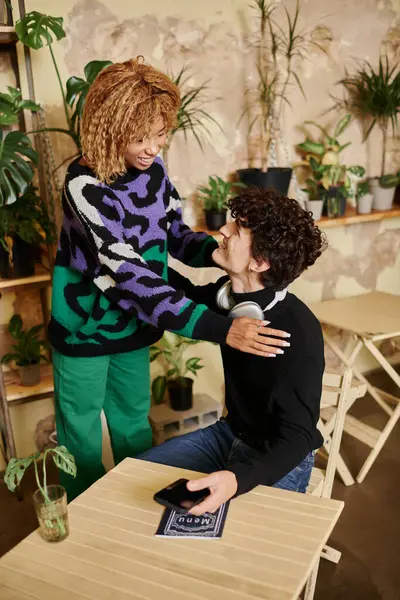 Mujer afroamericana feliz con el pelo rizado abrazando a hombre joven con teléfono inteligente en la cafetería vegana - foto de stock