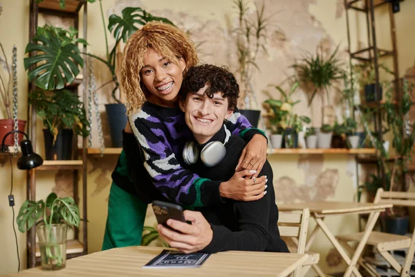Cheerful african american woman with curly hair embracing young man with  smartphone in vegan cafe — Stock Photo