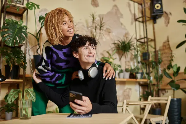 Femme américaine africaine positive avec les cheveux bouclés embrassant jeune homme avec smartphone dans un café végétalien — Photo de stock