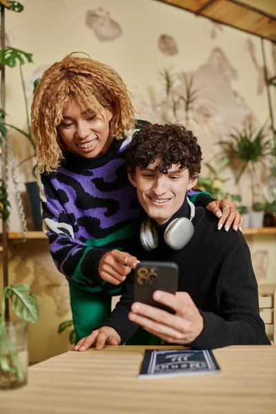 Mulher americana africana feliz com cabelo encaracolado abraçando jovem e olhando para o smartphone no café — Fotografia de Stock