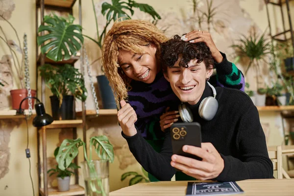 Весела афроамериканська жінка з кучерявим волоссям, що обіймає молодого чоловіка зі смартфоном у веганському кафе — стокове фото