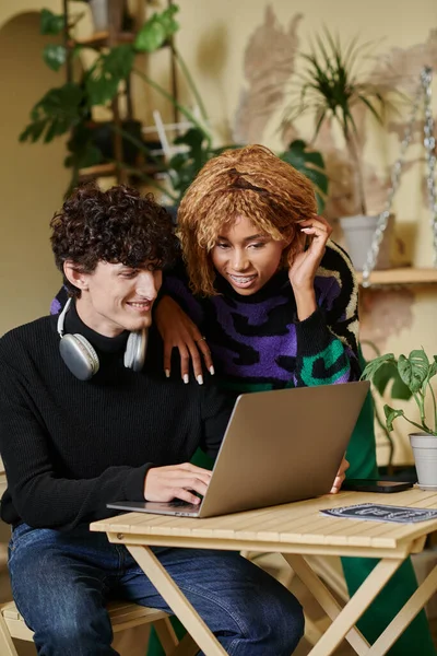 Mujer afroamericana feliz con frenos de pie cerca de novio y mirando a la computadora portátil en la cafetería - foto de stock