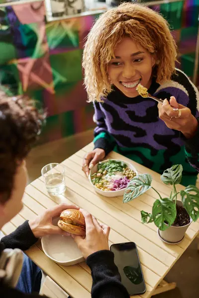 Щаслива афроамериканка в брекетах їсть смачний салат біля чоловіка з бургер у веганському кафе — стокове фото