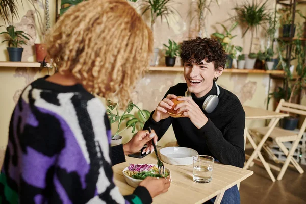 Happy curly man eating tofu burger near african american girlfriend enjoying salad in vegan cafe — Stock Photo
