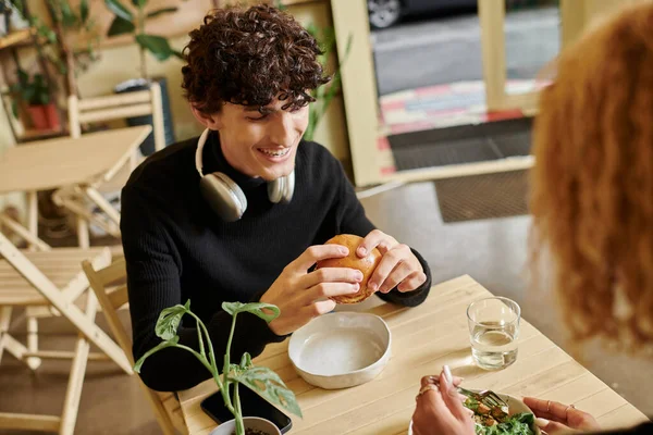 Cheerful and curly young man eating tofu burger near  girlfriend enjoying her meal in vegan cafe — Stock Photo