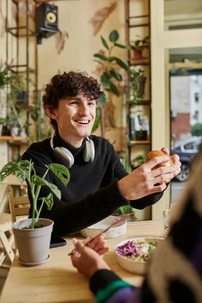 Fröhlicher und lockiger junger Mann isst Tofu-Burger in der Nähe seiner Freundin und genießt ihr Essen im veganen Café — Stockfoto