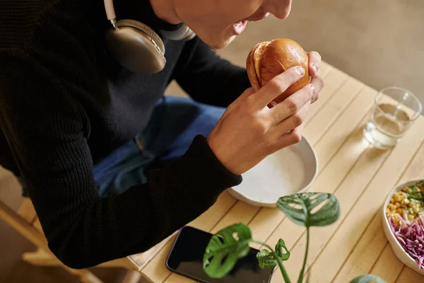 Geschnittenes Foto von jungem Mann, der Tofu-Burger in der Nähe von Smartphone und Salatschüssel in veganem Café isst — Stockfoto
