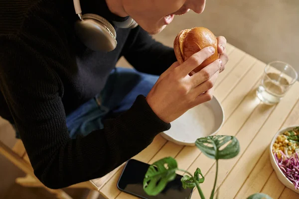 Cropped photo of young man eating tofu burger near smartphone and salad bowl in vegan cafe — Stock Photo