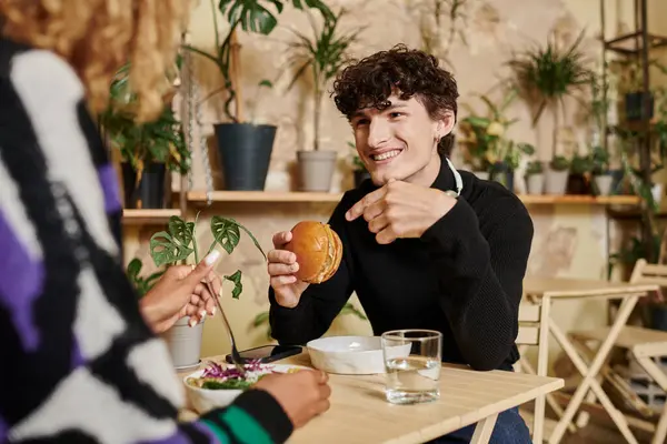 Alegre joven en auriculares sosteniendo hamburguesa de tofu y mirando a la mujer afroamericana en la cafetería - foto de stock