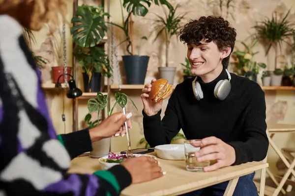 Hombre joven feliz en los auriculares mirando hamburguesa de tofu cerca de la mujer afroamericana feliz en la cafetería vegana - foto de stock