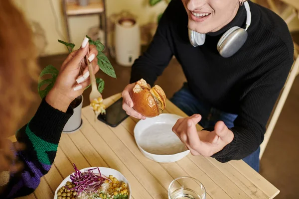 Plan recadré de l'homme heureux dans les écouteurs sans fil manger hamburger tofu végétalien près de la femme dans le café — Photo de stock
