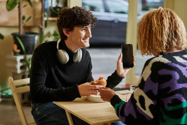 Happy curly man with wireless headphones holding tofu burger and smartphone in vegan cafe — Stock Photo