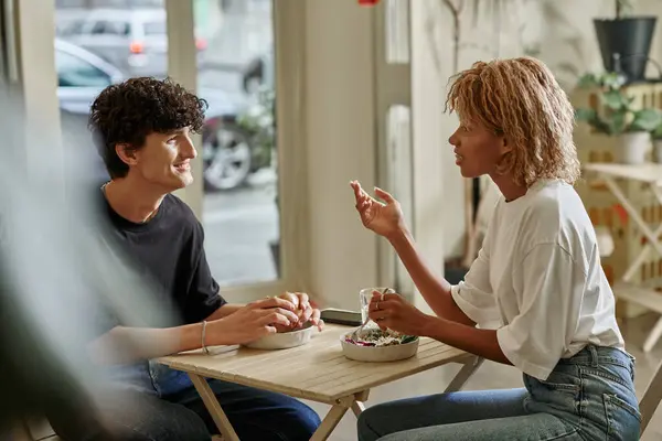 Happy curly man holding tofu burger and talking to african american young woman in vegan cafe — Stock Photo